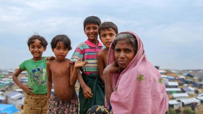 Cover Page - CCCM Case Studies 2016 - 2019. Four children stand smiling behind a woman seated on a hill. They are in Cox's Bazar